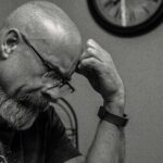 Black and white portrait of a thoughtful bald man indoors, capturing a moment of reflection with a wall clock in the background.
